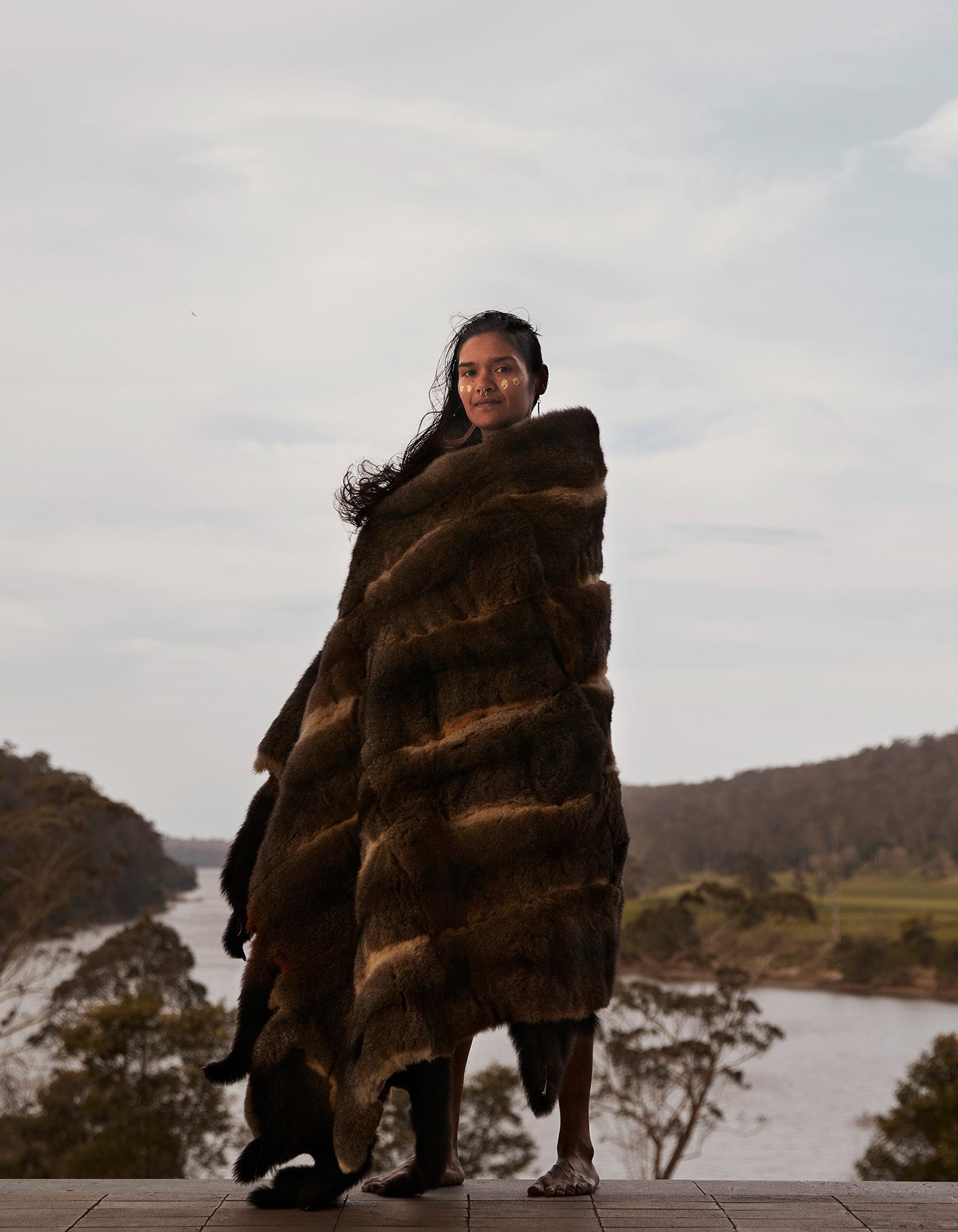 A women, standing in front of a rural landscape, wearing a Possum Skin Cloak.